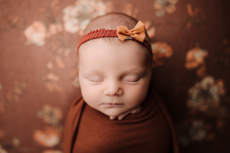 Newborn Photographer, a baby girl has a headband with bow, wrapped in a blanket near floral wall paper, all in a burnt orange color