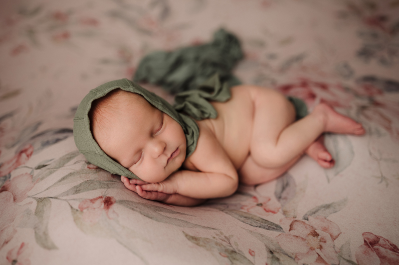 Newborn Photographer, a baby lays sleeping with a small hood on her head, she lays on a floral comforter