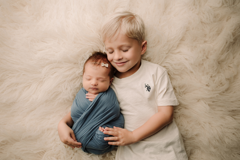 Newborn Photographer, a young boy lays beside his newborn baby sister, he smiles