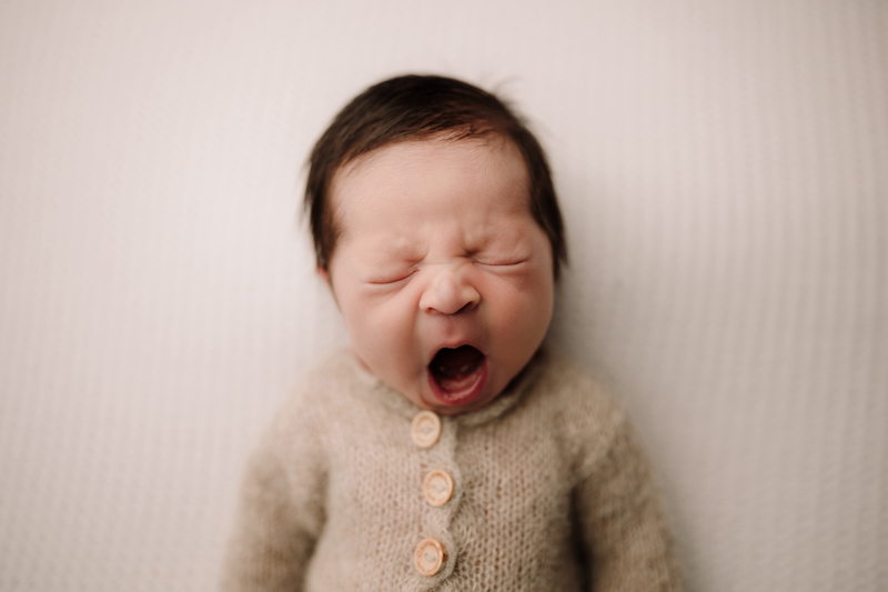 newborn photographer, a baby yawns with lots of expression, the baby is wearing a wool sweater with buttons